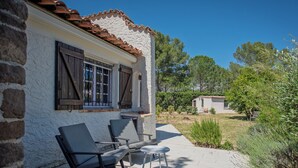A patio with two chairs and a table, a white building with a tiled roof, and a small shed in the background.