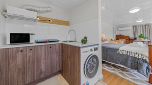 A compact hotel room with a kitchenette, a washing machine, a bed with a grey bedspread, and a small bedside table. - Convenient spot Gosford /near M1HW,CBD,Hospital,Stadium,water view,fishing,pet (West Gosford)
