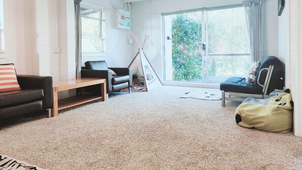 A spacious living room with a sofa, a chair, a bench, a window, and a sliding glass door leading to a garden. - 2 bedroom cosy guest house in North shore (Auckland)