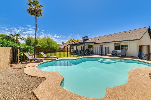 A swimming pool with a curved edge, surrounded by a fence, and a house with a covered patio area.