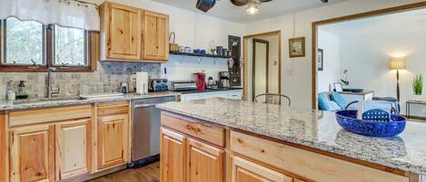 A kitchen with wooden cabinets, granite countertops, and a large island. There are pots hanging from the ceiling, a sink, and a stove.