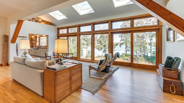 A spacious living room with a high ceiling, skylights, large windows, a sofa, a chair, a wooden cabinet, and a small kitchen area. - Chic Addison Home w/ Fire Pit on 8 Private Acres! (Addison)