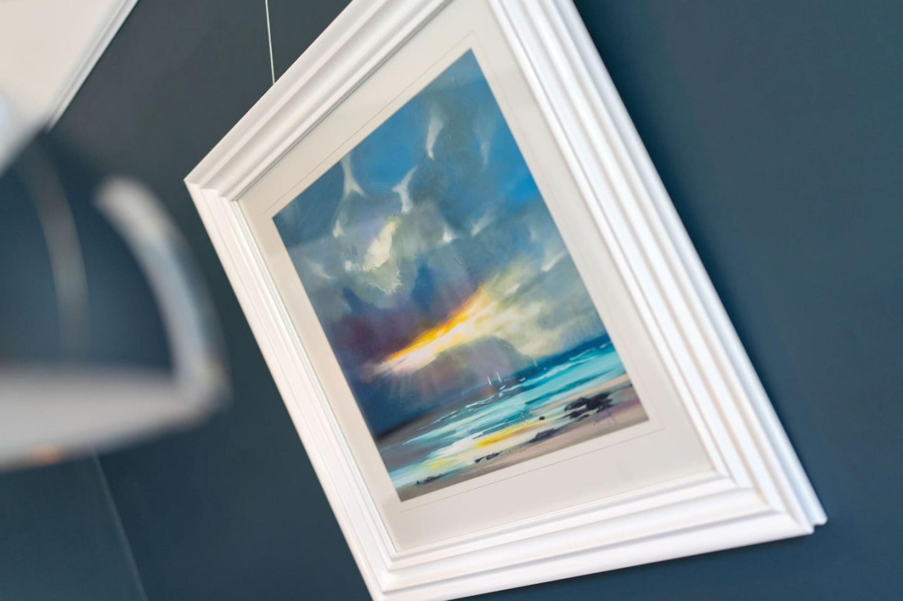 A framed painting of a seascape with a cloudy sky and a sunset, hanging on a dark wall.
