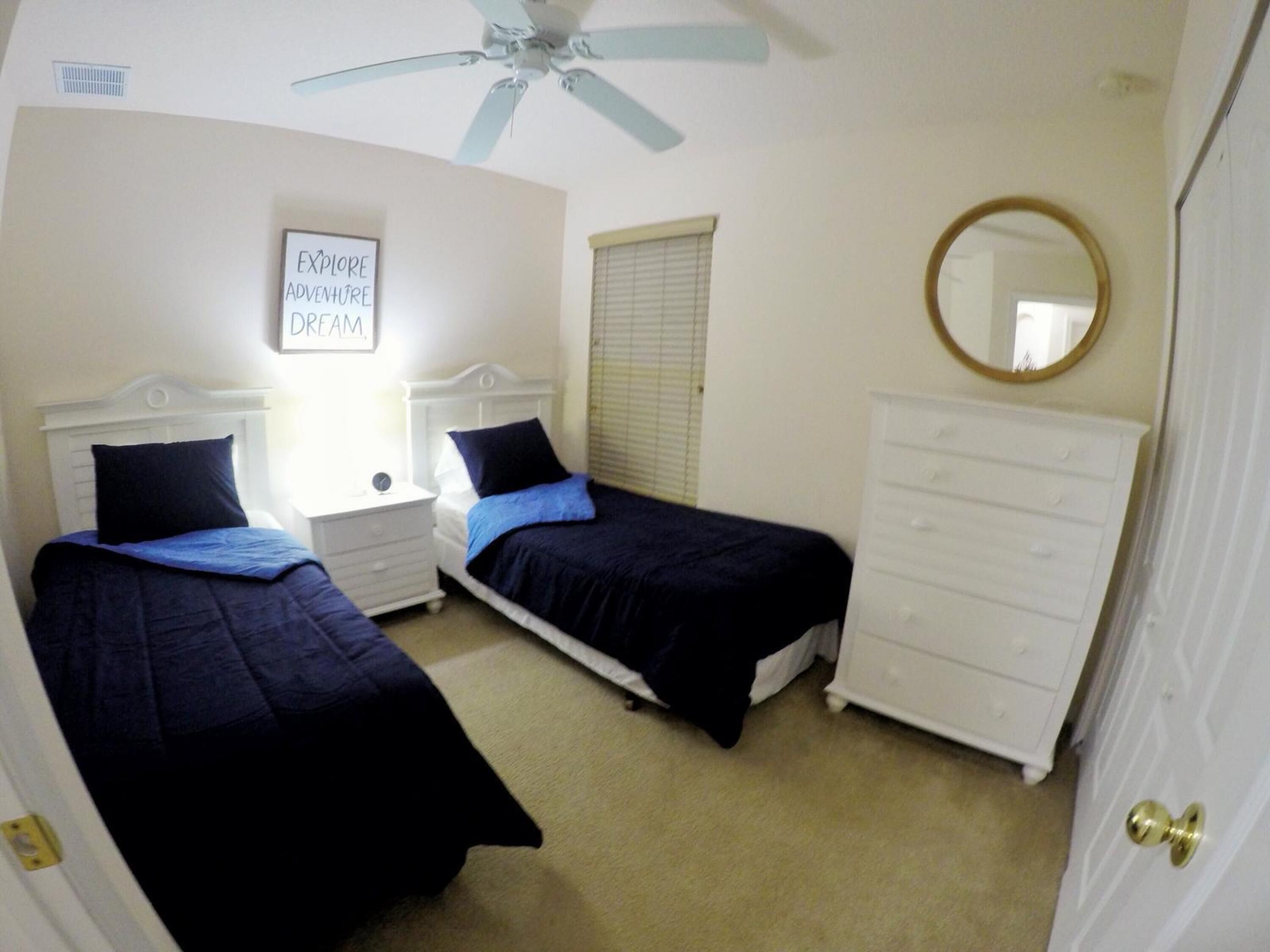 A bedroom with two beds, a ceiling fan, a bedside table, a dresser, a window with blinds, and a mirror.