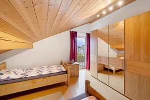 Ein Schlafzimmer mit schrägen Wänden, einem Bett, einem kleinen Tisch, einem Spiegel und Blick durch ein Fenster mit Vorhängen.
