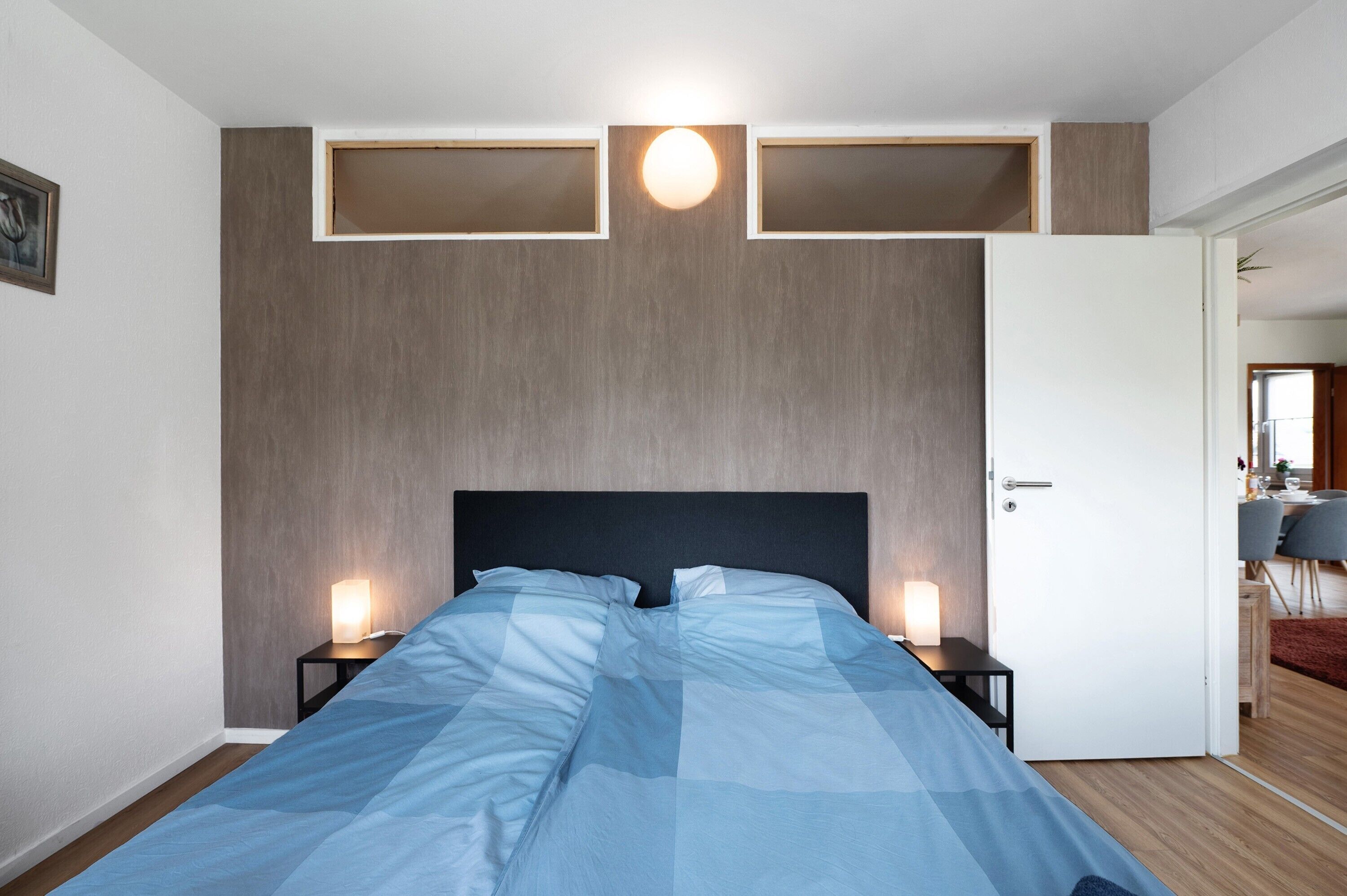 A bedroom with a bed, bedside tables, a nightstand, a door, and a wall-mounted light fixture.