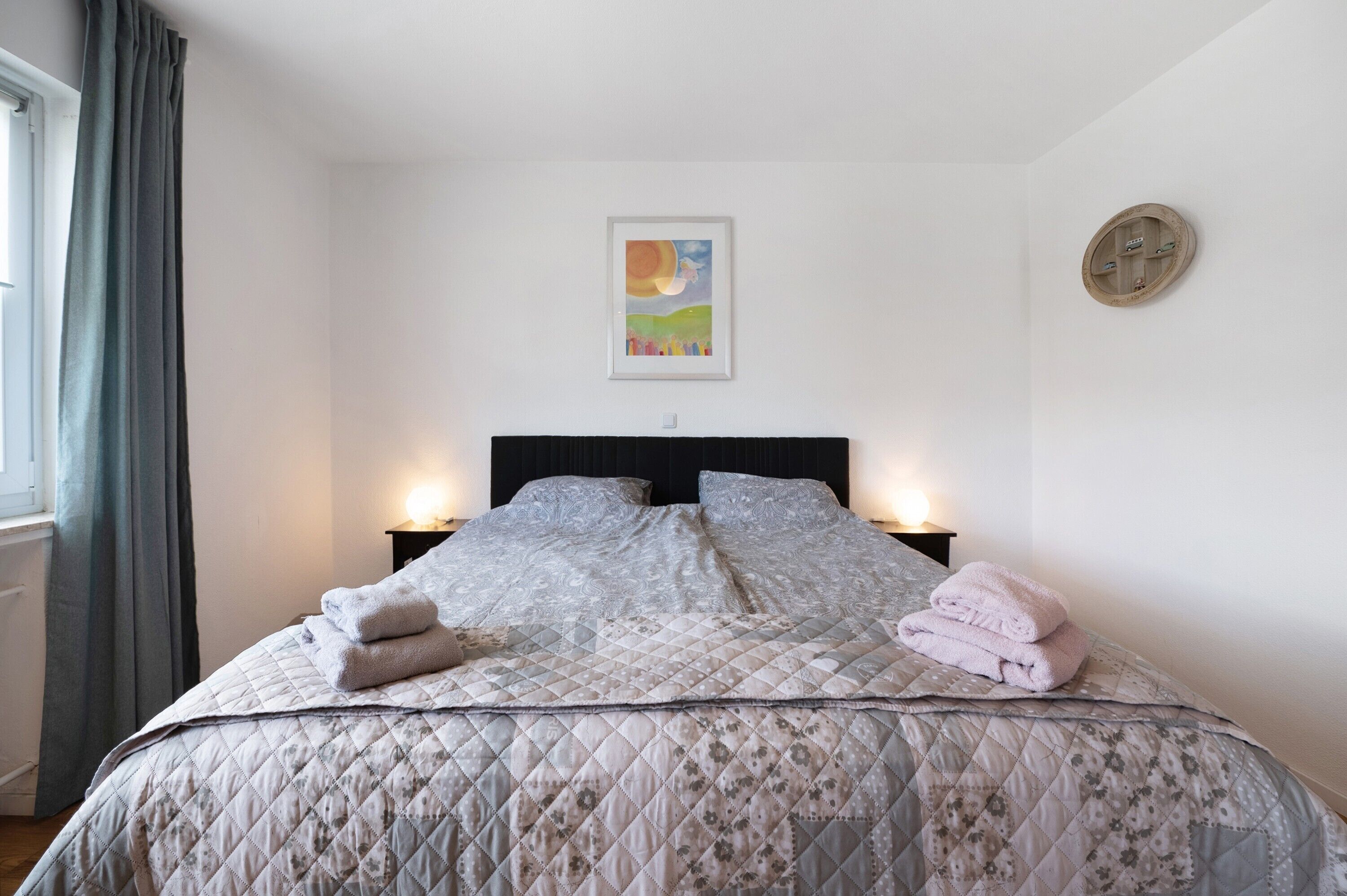 A bedroom with a bed, two bedside tables, a painting on the wall, and a round mirror.