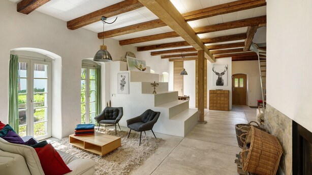 Ein geräumiges Wohnzimmer mit hoher Decke, sichtbaren Holzbalken, einem Kamin, einer Treppe und Blick durch große Fenster.