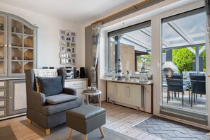 A modern kitchen with a grey sofa, a dining area with outdoor seating, and a large window with a view of greenery.