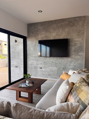 Una sala de estar moderna con un ventanal grande, un televisor de pantalla plana montado en una pared con textura, una mesa de centro de madera y un sofá con cojines estampados.