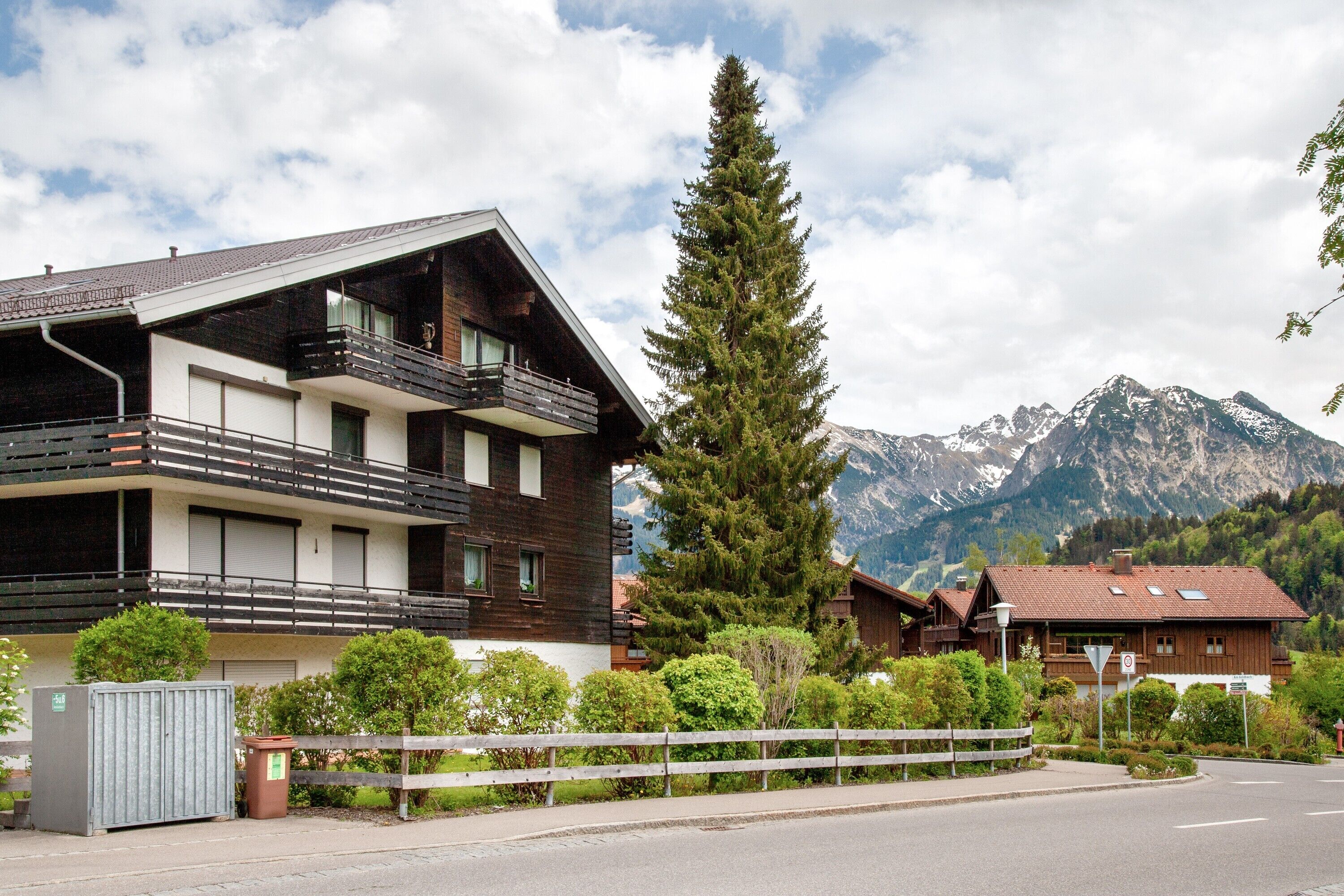 Un bâtiment moderne à deux étages, doté de balcons, d’un grand arbre à feuilles persistantes et surplombant un paysage montagneux.