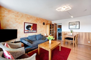 A living room with a sofa, a coffee table, wicker chairs, a red rug, a TV, a wooden cabinet, and a dining area with a table and chairs. - Apartment 'Bergrose' with Hörnerdörfer Mountain Cable Car Pass, Pool & Sauna Included (Obermaiselstein)