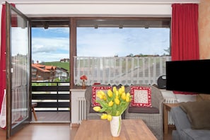 A living room with a wooden table, a sofa, a TV, a window with a view of houses, and a vase with flowers. - Apartment 'Bergrose' with Hörnerdörfer Mountain Cable Car Pass, Pool & Sauna Included (Obermaiselstein)
