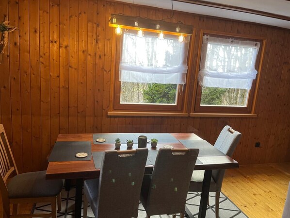 A wooden-paneled room with a dining table, chairs, and two windows with curtains.