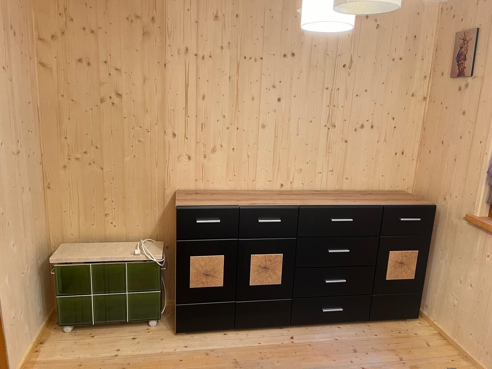 Ett rum med träväggar, ett svart skåp med lådor, ett grönt skåp och en vit lampa.