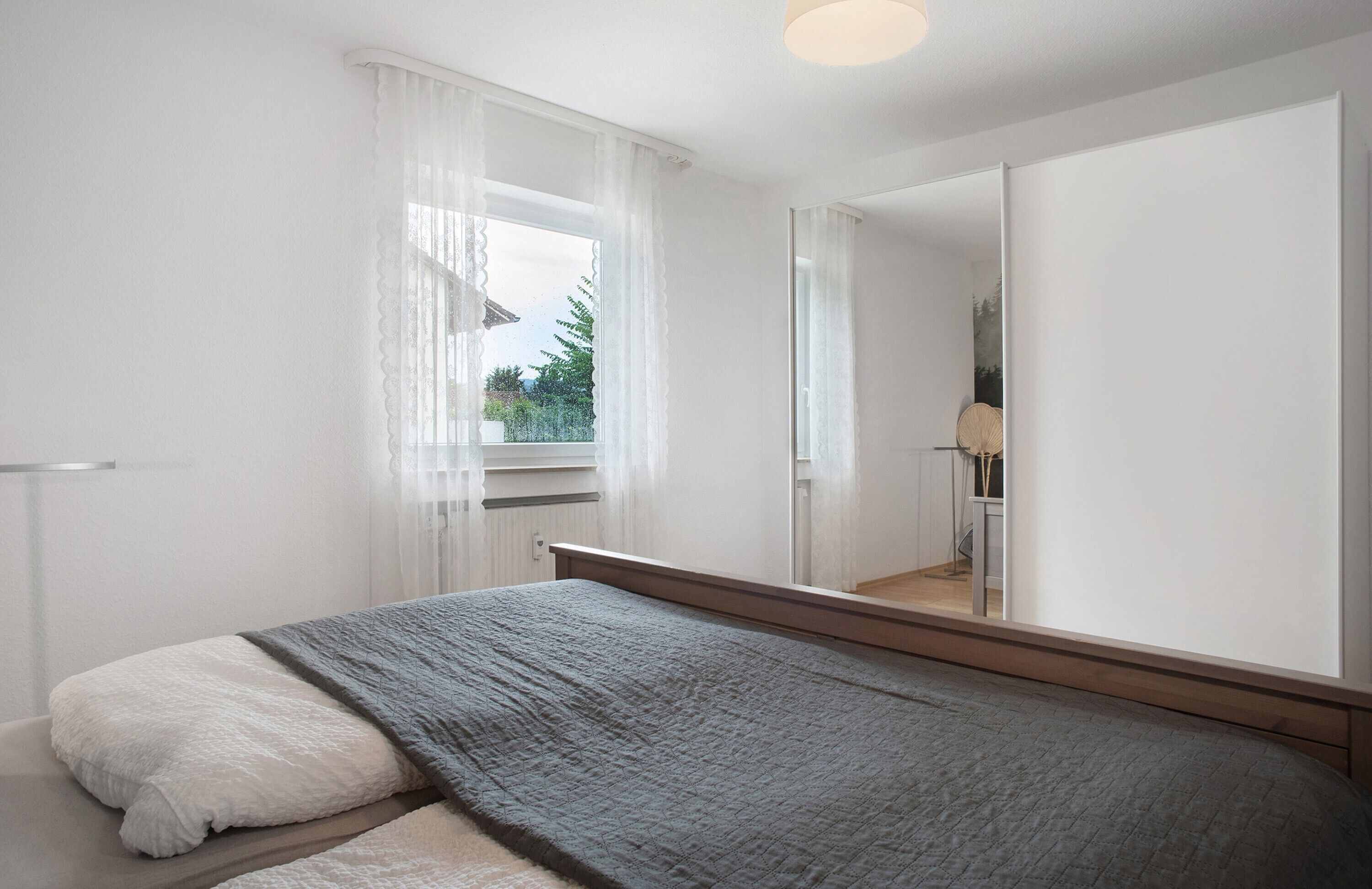 A bedroom with a bed, a window with sheer curtains, a mirror, and a view of greenery outside.