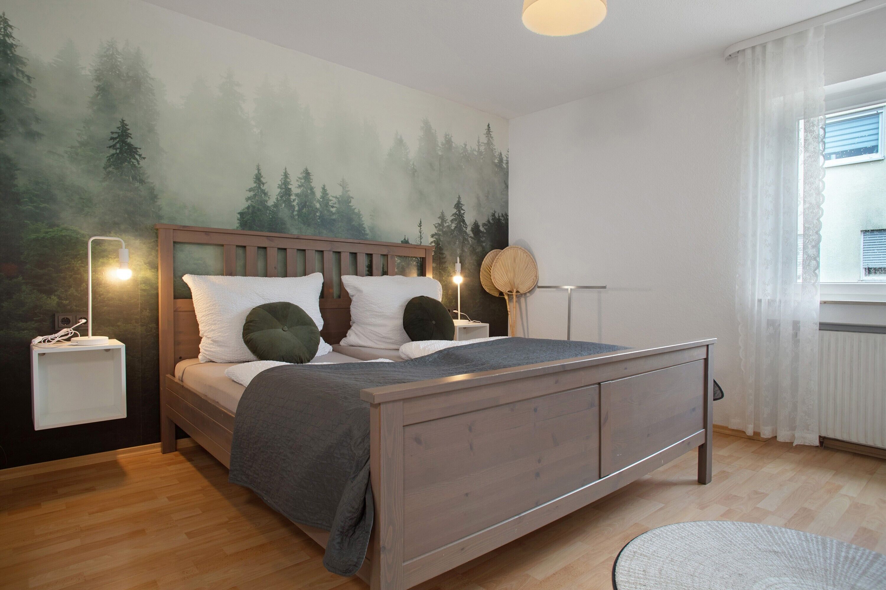 Un dormitorio con cama, mesitas de noche, una lámpara de pared, una mesita auxiliar y una ventana con cortinas.