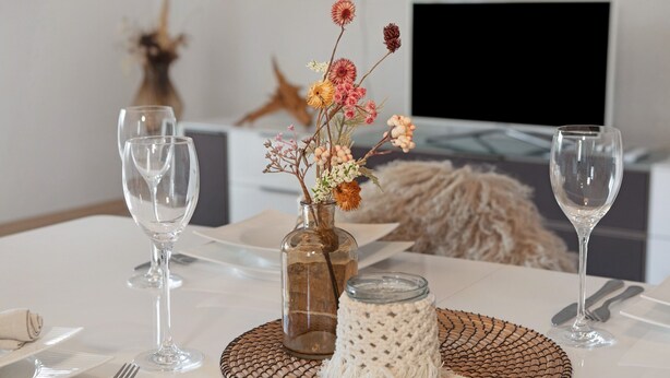 Una mesa de comedor dispuesta con cristalería transparente, un jarrón decorativo con flores secas y un mantel individual tejido.