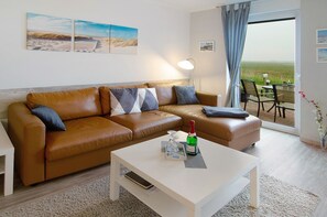Un salon moderne avec un canapé en cuir marron, une table basse blanche et une vue sur un champ verdoyant, visible à travers une porte vitrée.