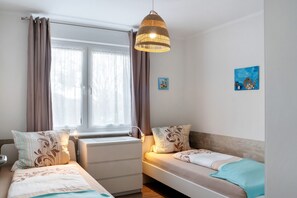 A bedroom with two beds, a window with curtains, a hanging light fixture, and wall art.