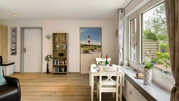 A room with a wooden floor, a white dining table with chairs, a bookshelf, a painting of a lighthouse, and a window with curtains.