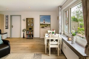 Una habitación con piso de madera, una mesa de comedor blanca con sillas, una estantería, un cuadro de un faro y una ventana con cortinas.