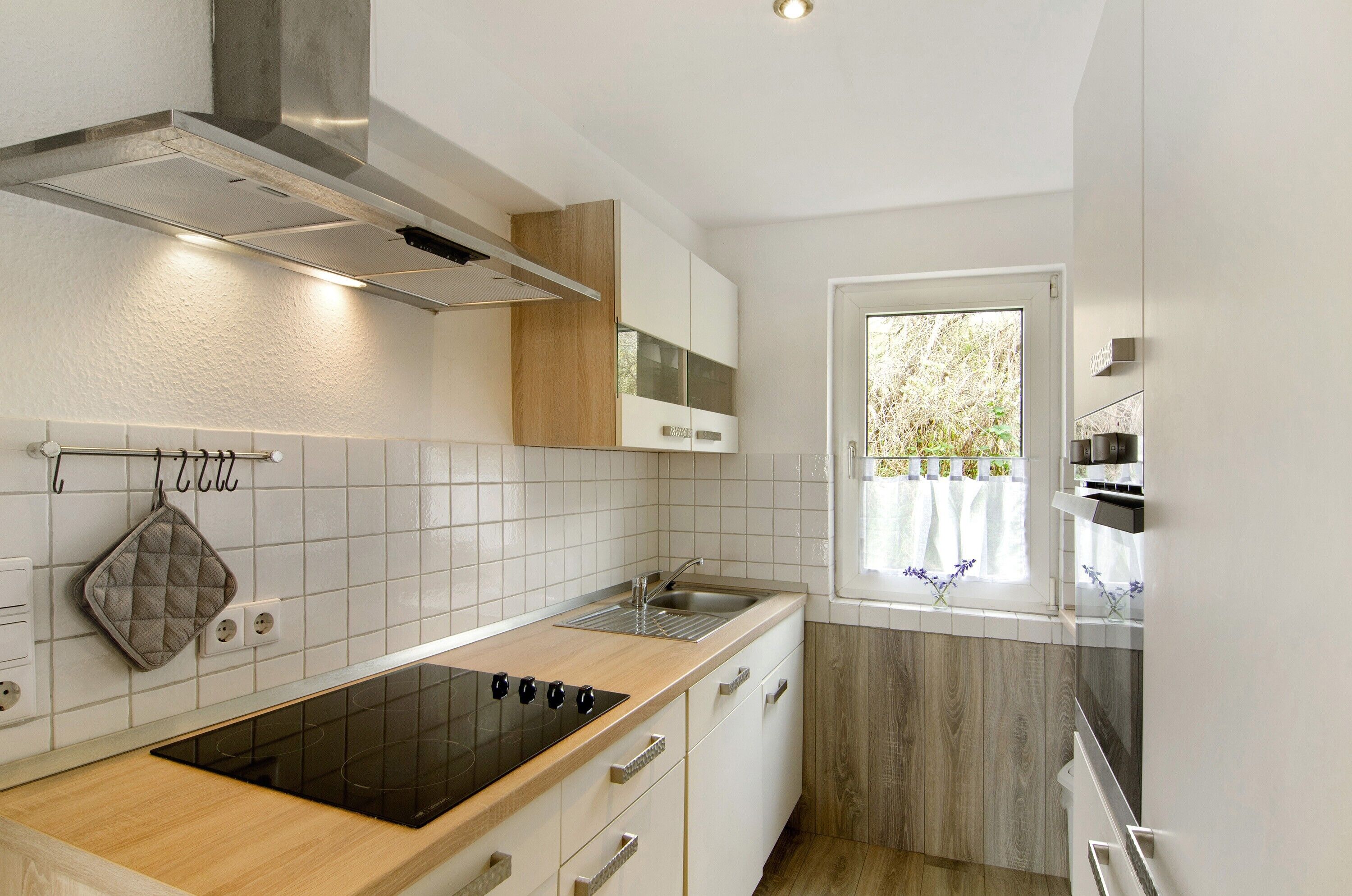 A modern kitchen with white tiles, wooden cabinets, a built-in oven, and a window with a view of greenery.