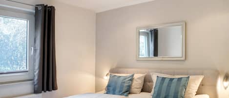 A bedroom with two beds, a window with curtains, a mirror, and a bedside lamp.