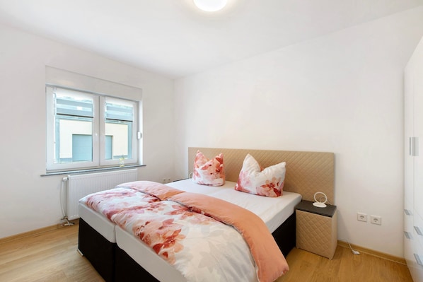 A bedroom with a bed, a bedside table, a nightstand, a window, and a radiator.