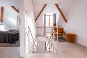 Une chambre de style loft moderne, avec un plafond en pente, un escalier, un lit et un coin salon comprenant un fauteuil et un pouf.