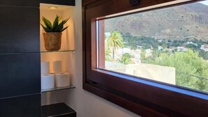 A room with a large window, a shelf with a plant and toilet paper, and a view of a hillside with buildings. - Natural beauty at Sol Caldera de Temisas (Temisas)