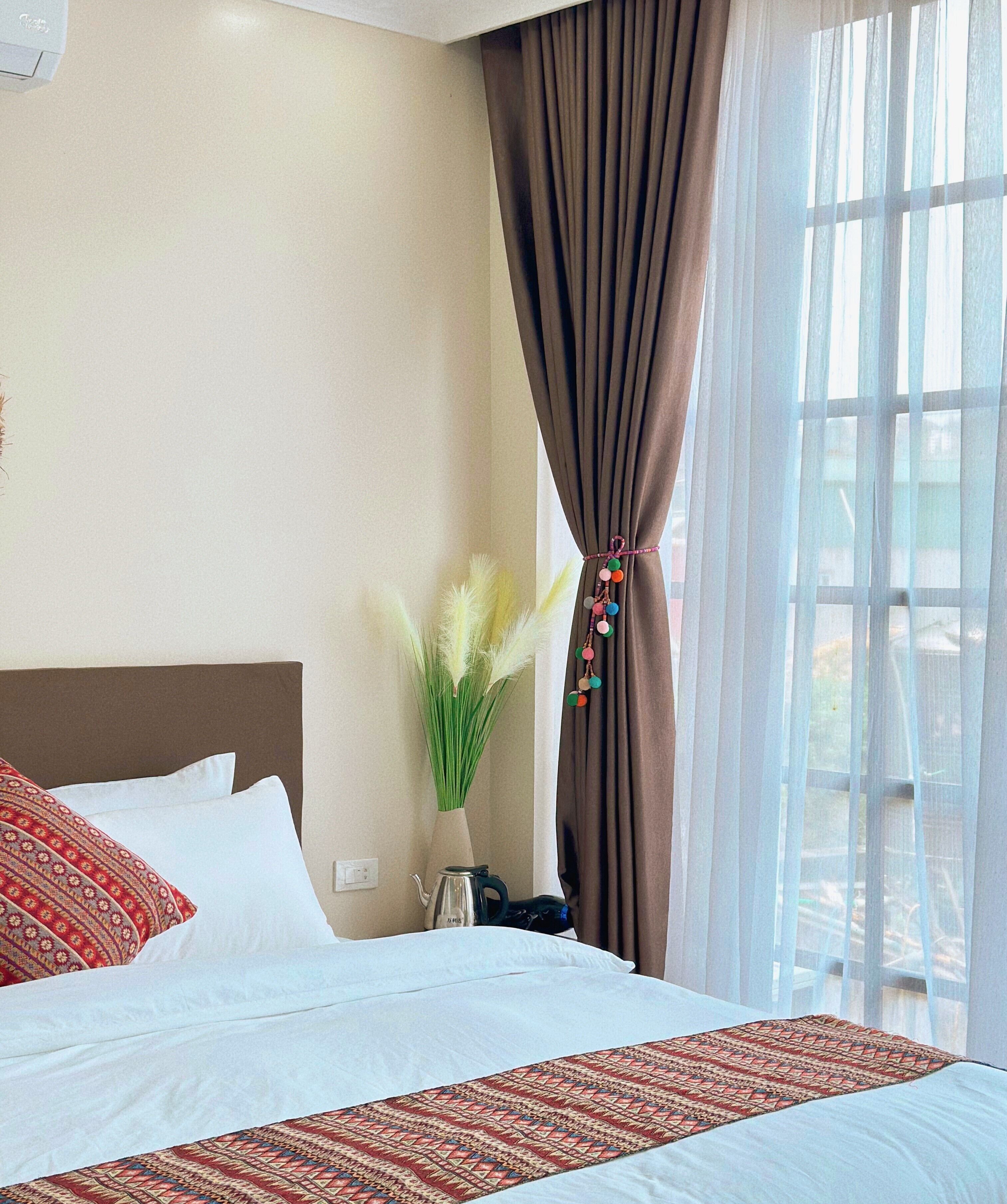 A hotel room with a bed, a vase with green plants, a kettle, and a window with curtains.