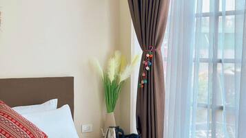 A hotel room with a bed, a vase with green plants, a kettle, and a window with curtains.