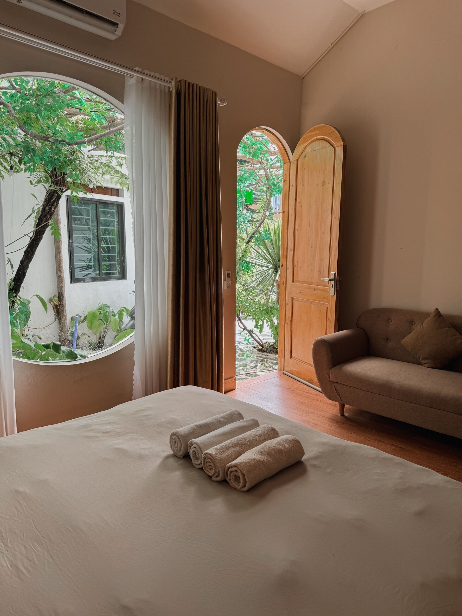 A bedroom with a bed, a wooden door, a window with curtains, and a view of greenery outside.