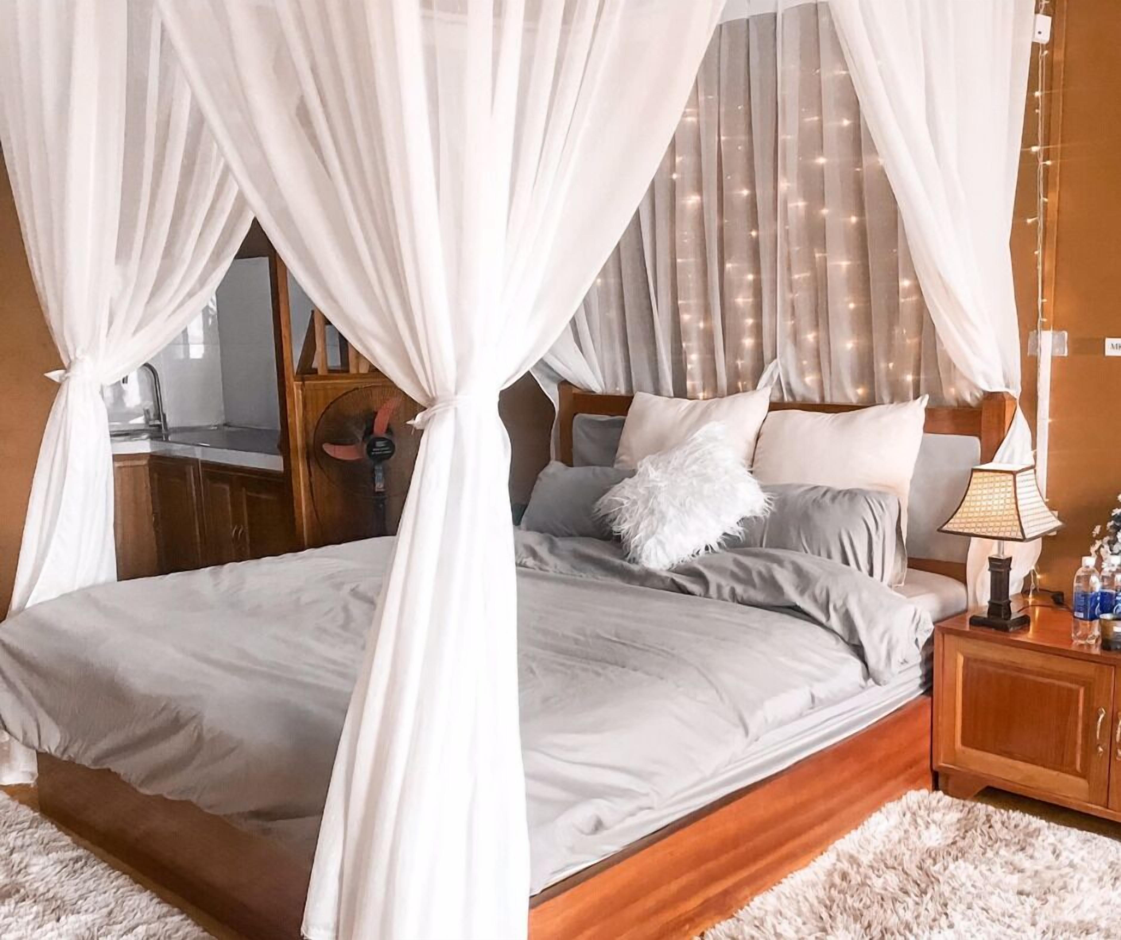 A bedroom with a four-poster bed, white bedding, a fluffy pillow, a bedside table with a lamp, and a wooden nightstand.