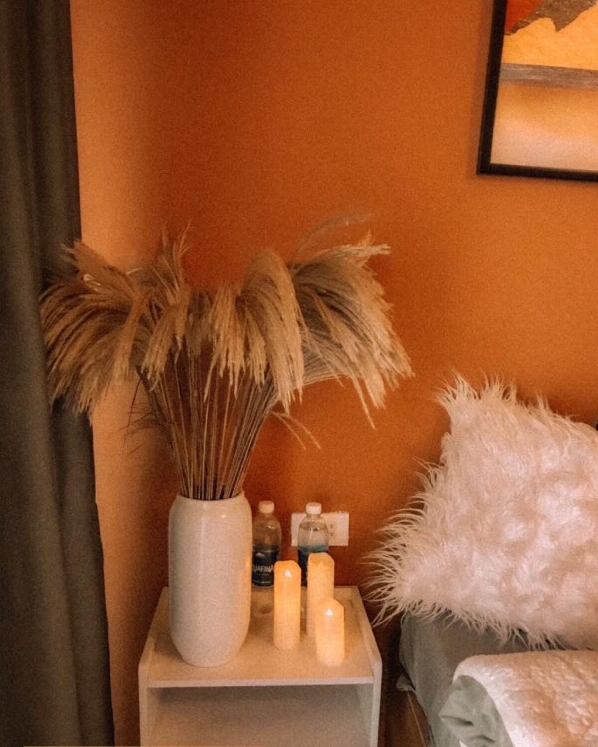 A bedside table with a vase of dried pampas grass, three candles, and a bottle of water.