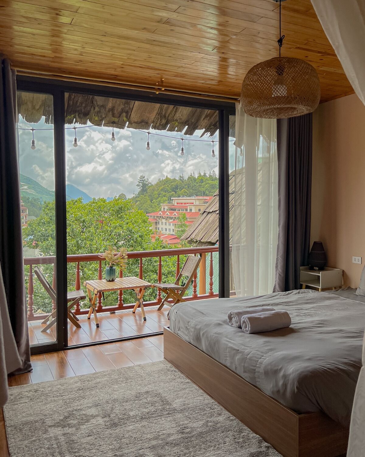 Un dormitorio con una cama grande, techo de madera, balcón con mesa y sillas, y vista a colinas verdes y edificios.