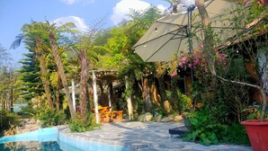 Zona de piscina con terraza de piedra, rodeada de exuberante vegetación y una pérgola con luces colgantes.