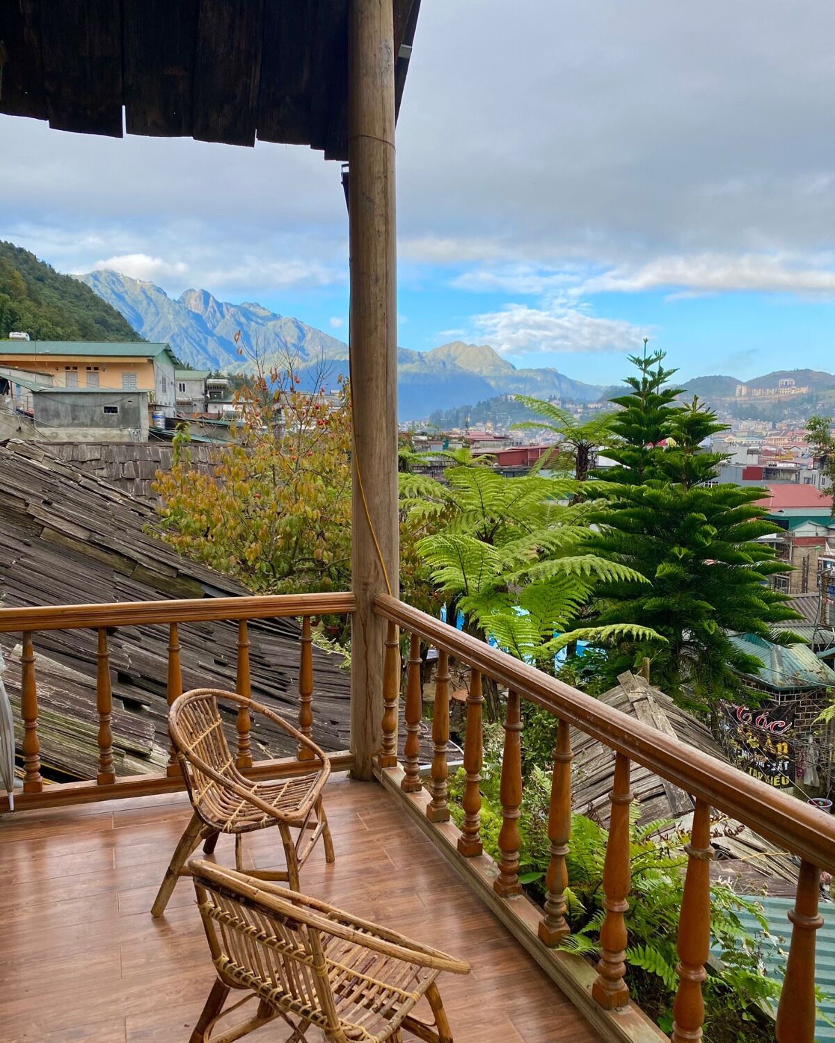 Ein Balkon mit Korbstühlen, einem Holgitter und Blick auf eine gebirgige Landschaft mit Gebäuden in der Ferne.