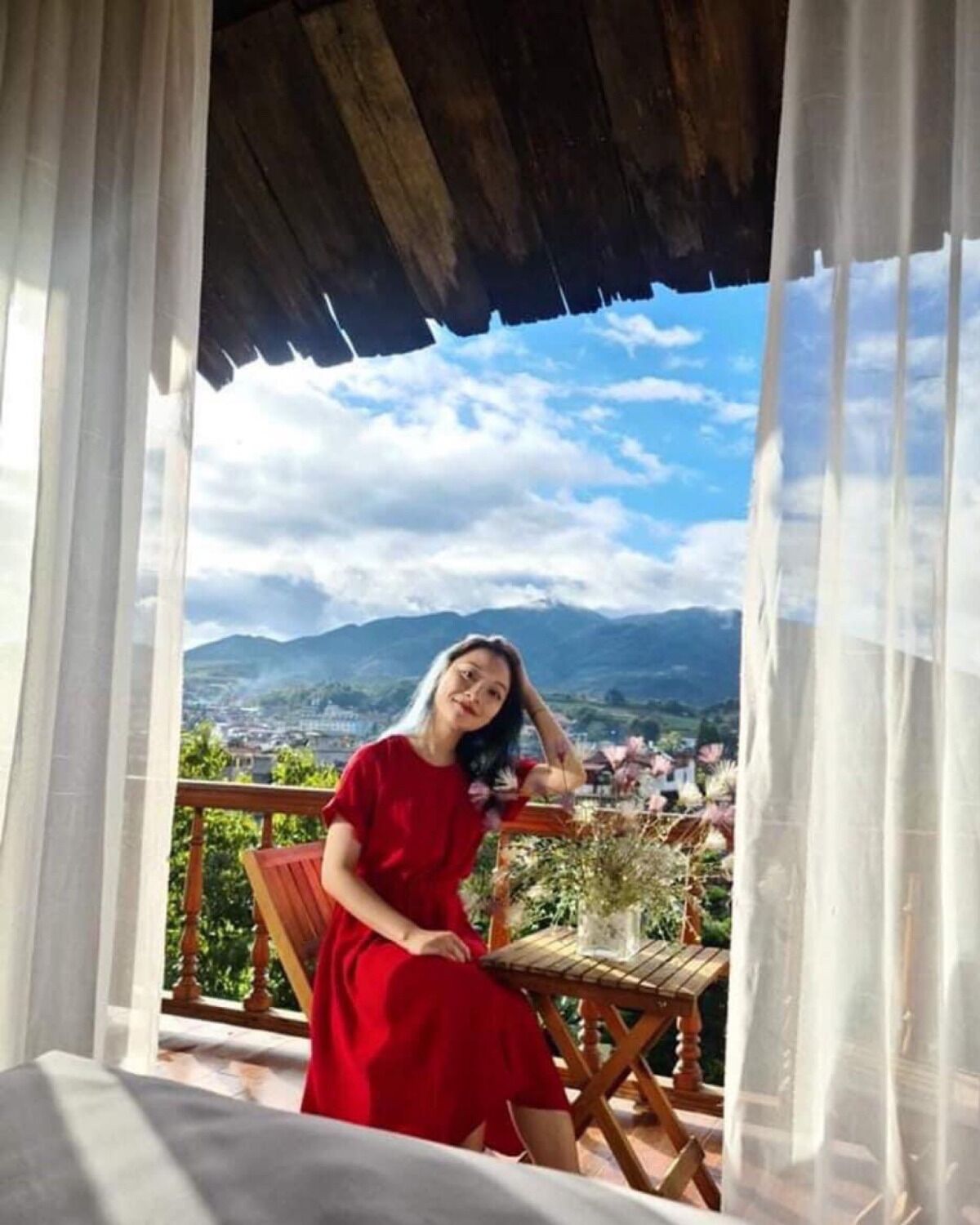 A person in a red dress sitting on a wooden chair on a balcony with a view of mountains and a town.