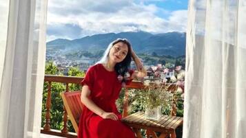 A person in a red dress sitting on a wooden chair on a balcony with a view of mountains and a town.