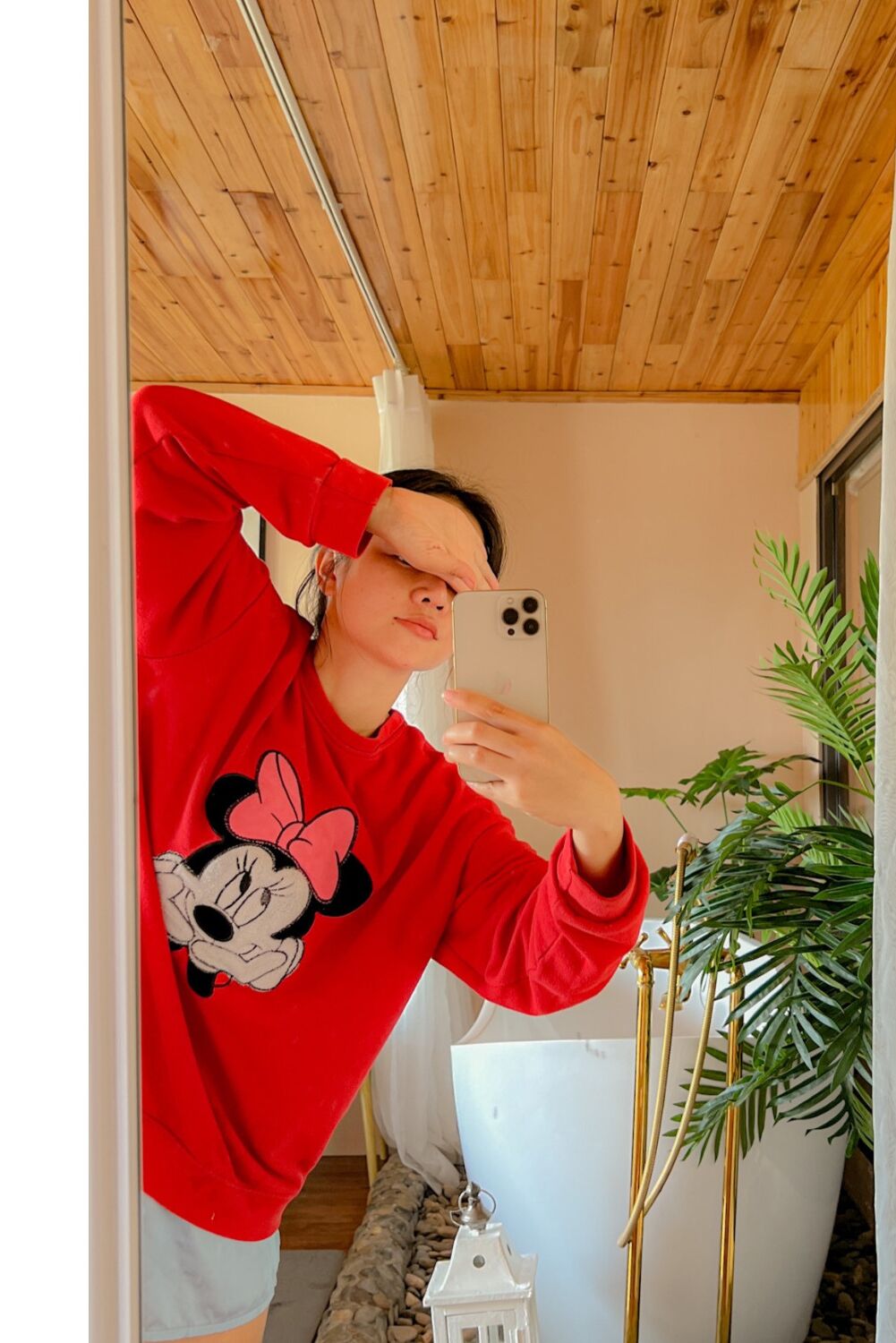 A person taking a selfie in a room with wooden ceiling, a white wall, and a potted plant.