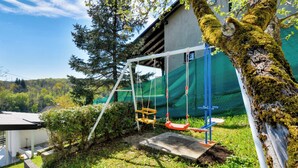 A swing set in a garden area with a tree covered in moss, a concrete structure, and a building in the background. - Haven Near The River (Duga Resa)