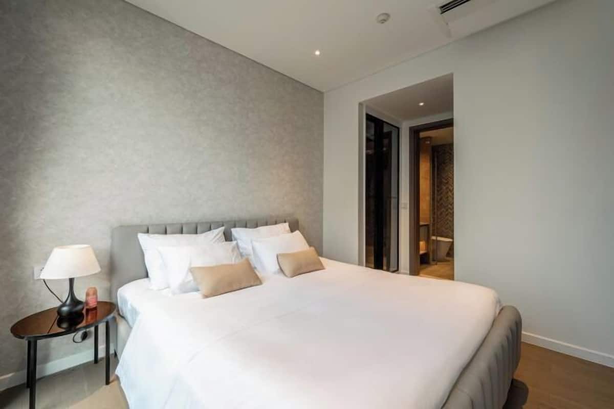 Una habitación de hotel con una cama grande, una mesita de noche con lámpara y una puerta que da a otra habitación.