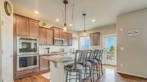 A modern kitchen with wooden cabinets, stainless steel appliances, a central island with bar stools, and a view to the outside. - Mountain Breeze! Fire Pit, Air Hockey & Views! (Monument)