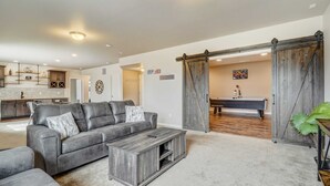 A spacious living room with a gray leather sofa, a wooden coffee table, and a pool table. A kitchen with modern appliances and a dining area with a large wooden table are visible in the background. - Mountain Breeze! Fire Pit, Air Hockey & Views! (Monument)