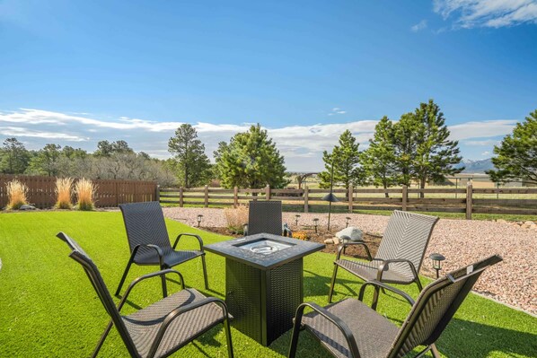An outdoor patio with a square table and four chairs, surrounded by a wooden fence and trees. - Mountain Breeze! Fire Pit, Air Hockey & Views! (Monument)