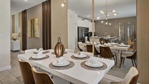 A modern dining area with a long table set for four, a chandelier, and a kitchen in the background.