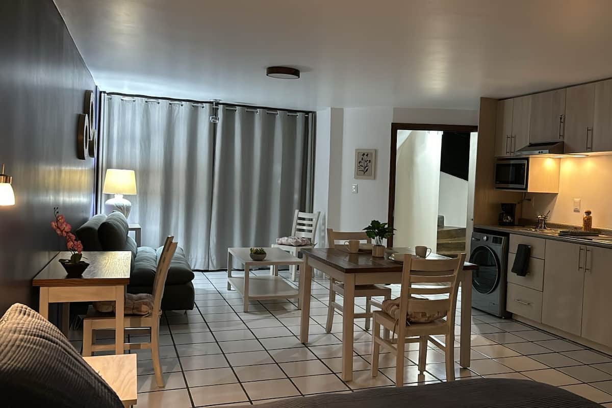 Uma cozinha moderna com eletrodomésticos de aço inoxidável, uma área de jantar com mesa e cadeiras, e uma sala de estar com sofá e abajur.