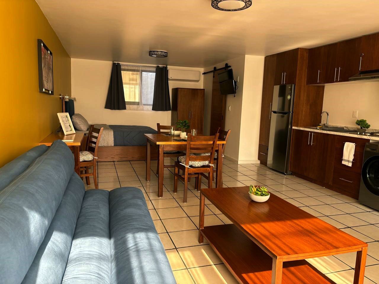 A living room with a sofa, dining table, chairs, and a kitchen area with cabinets and appliances.